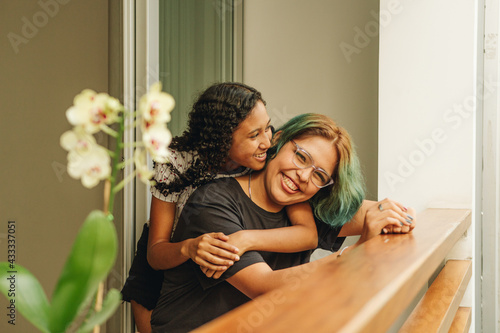 Pareja de jóvenes lesbianas expresan su cariño mediante un abrazo en su casa mientras una mira hacia la calle  © Gerardo