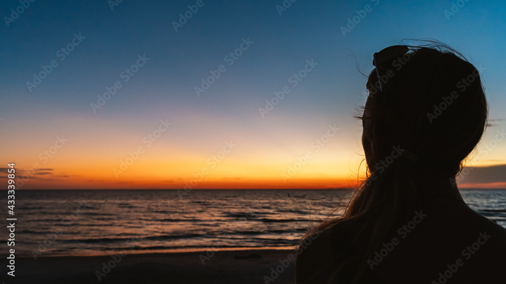 Woman watches sunset over ocean beach in silhouette from behind