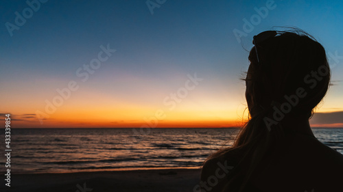 Woman watches sunset over ocean beach in silhouette from behind