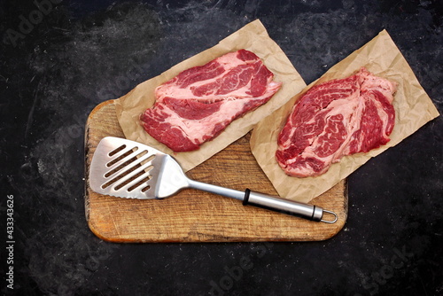 Raw Marbled Loin Beef Steaks and Grill Tools On Wooden Cutting Board. Beef Steaks On Spatula Ready for BBQ or Grilling, Overhead View. Raw Striploin Marbled Beef Steaks on Black Background, Top View.