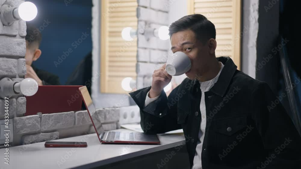 Portrait of satisfied Chinese young man posing in home office on break. Smiling handsome Asian manager sitting in bedroom at mirror with lights drinking coffee looking at camera with laptop aside Portrait of satisfied Chinese young man posing in home office on break. Smiling handsome Asian manager sitting in bedroom at mirror with lights drinking coffee looking at camera with laptop aside
