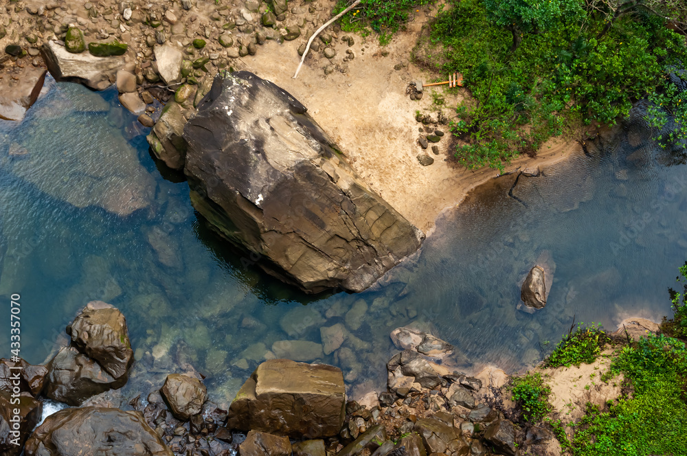 Closeup of the base of the Pinaisara waterfall seen from the top with beautiful natural pool, lush vegetation, rock formations, trail signage on the ground. Iriomote Island.