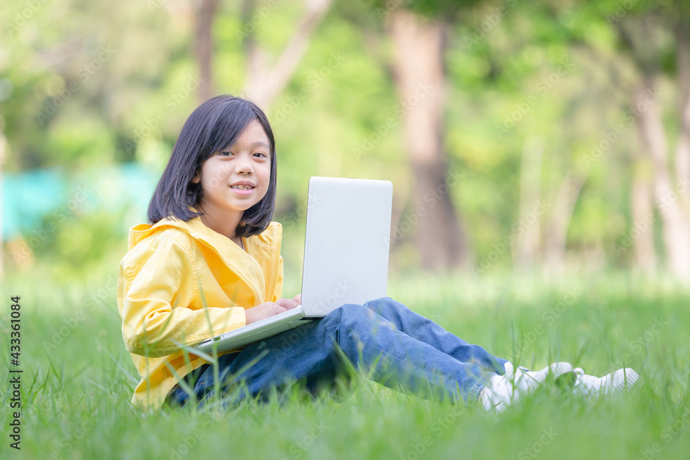 Asian kid girl sitting on the grass studying with laptop in the park, Outdoor learning concepts Asian kid girl sitting on the grass studying with laptop in the park, Outdoor learning concepts