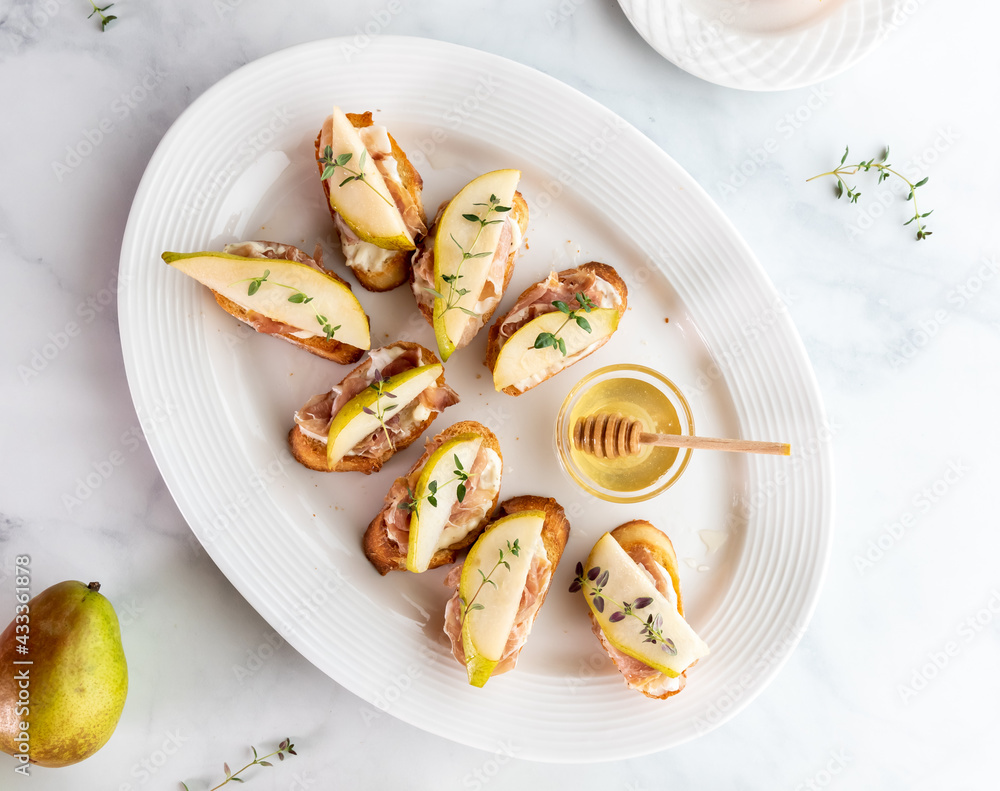 Top down view of a platter of crostini bites topped with sliced pear and prosciutto and thyme. Top down view of a platter of crostini bites topped with sliced pear and prosciutto and thyme.