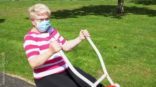 Senior blonde woman with face mask working out arms on park machine on sunny day. Elder lady training to keep fit and healthy outdoors