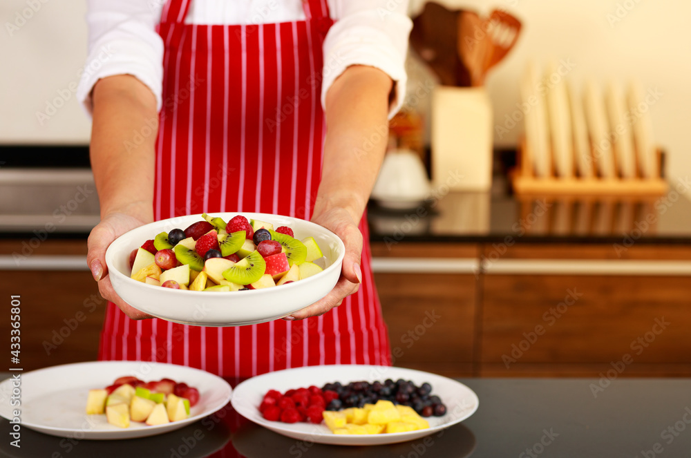 Closeup shot of tasty yummy sweet mixed fruit salad appetizer apple kiwi blueberry grape in white bowl on executive female chef hands who stand wears stripe red apron behind in background