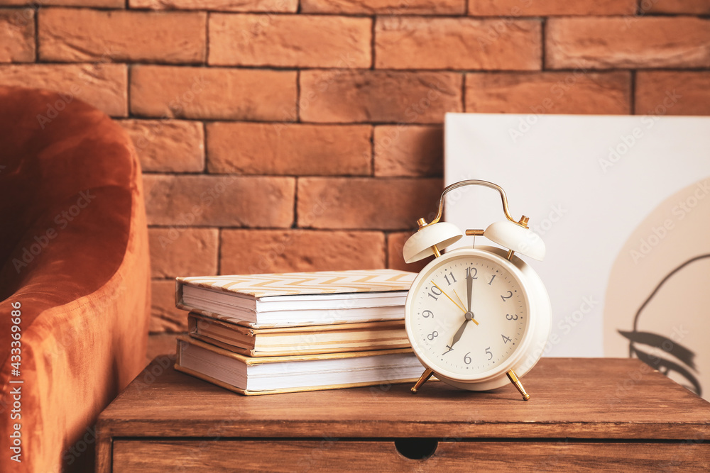 Table with alarm clock and books near brick wall in room Table with alarm clock and books near brick wall in room