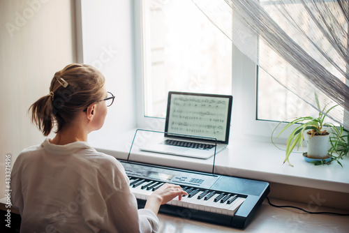 View from the back young woman plays a synthesizer, reading notes on a laptop screen. Independent learning to play the piano at home. Passion for music, hobbies, leisure, self-development.