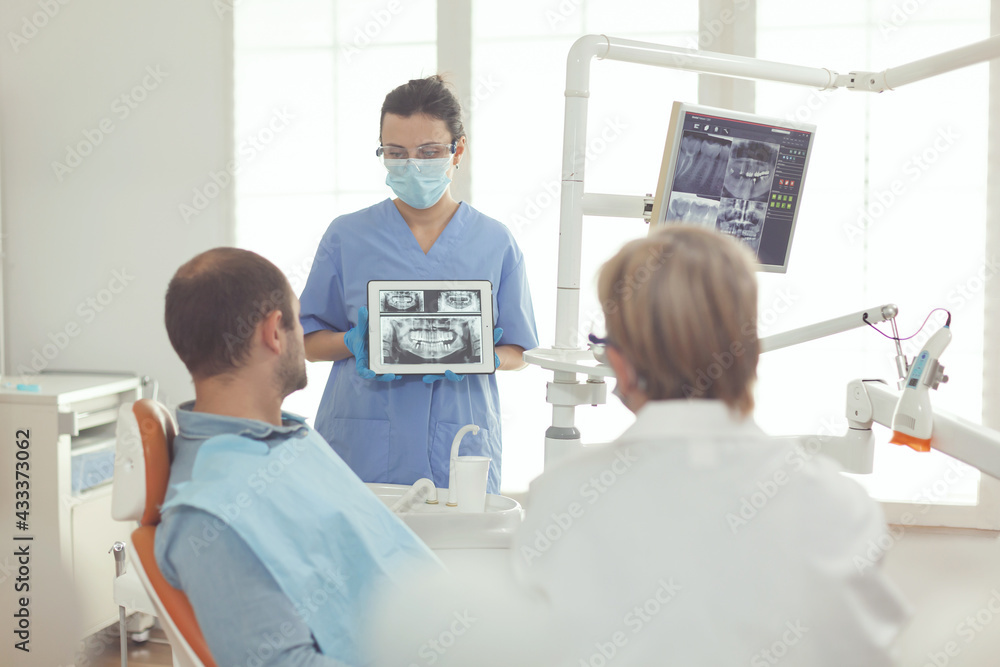 Senior somatology woman talking to sick man about tooth healthcare while sitting in dental chair. Orthodontist nurse explaining toothache pain using digital tablet with denture xray on screen Senior somatology woman talking to sick man about tooth healthcare while sitting in dental chair. Orthodontist nurse explaining toothache pain using digital tablet with denture xray on screen