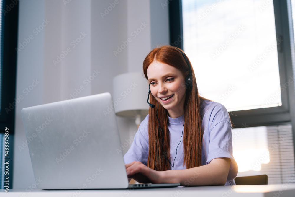 Close-up of happy young woman operator using headset and laptop during customer support at home office. Young female student communicating online by video call on background of window. Close-up of happy young woman operator using headset and laptop during customer support at home office. Young female student communicating online by video call on background of window.