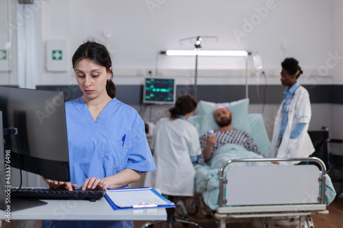 Woman asisstant typing medical recovery on computer while in background practitioner doctor monitoring sick man explaining disease symptom. Hospitalized patient having respiratory disorder photo