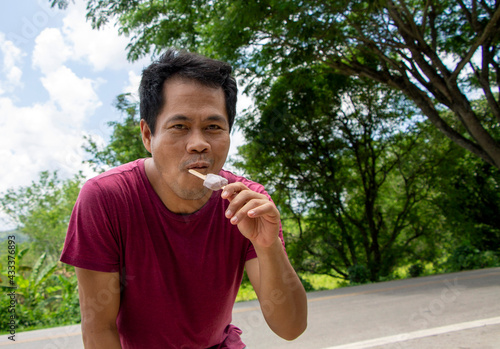 portrait of an Asian man dress casually standing smiling enjoys eating ice cream by a countryside road. Happy and healthy male concept