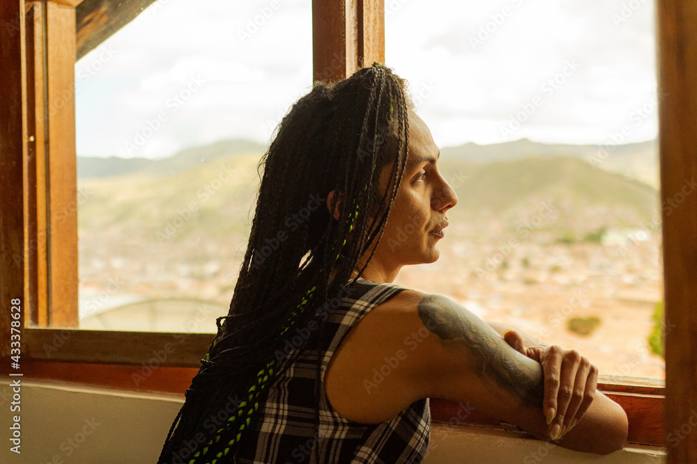 Mujer transgenero tatuada y con trenzas mirando el paisaje a través de su ventana  - obrazy, fototapety, plakaty 