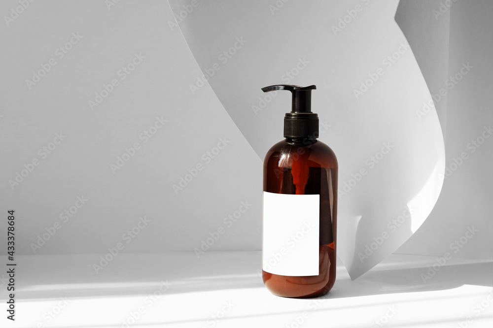 Brown bottle with dispenser for cream, shampoos, oils with blank label on white background with an unfolded sheet of paper. Solid background with copyspace and falling shadow from window