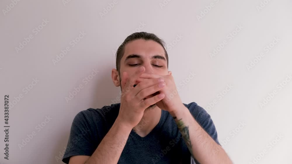 Young handsome Caucasian man with beard and ponytail sits in blue Tshirt on white background and plays harmonica completely immersed in melody. Playing compact musical instrument. Young handsome Caucasian man with beard and ponytail sits in blue Tshirt on white background and plays harmonica completely immersed in melody. Playing compact musical instrument.