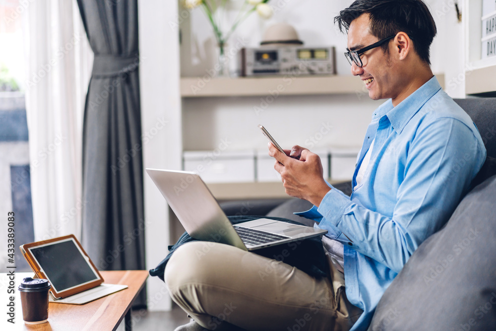 Young smiling asian man relaxing using laptop computer working and video conference meeting at home.Young creative man looking at screen typing message with smartphone.work from home concept Young smiling asian man relaxing using laptop computer working and video conference meeting at home.Young creative man looking at screen typing message with smartphone.work from home concept