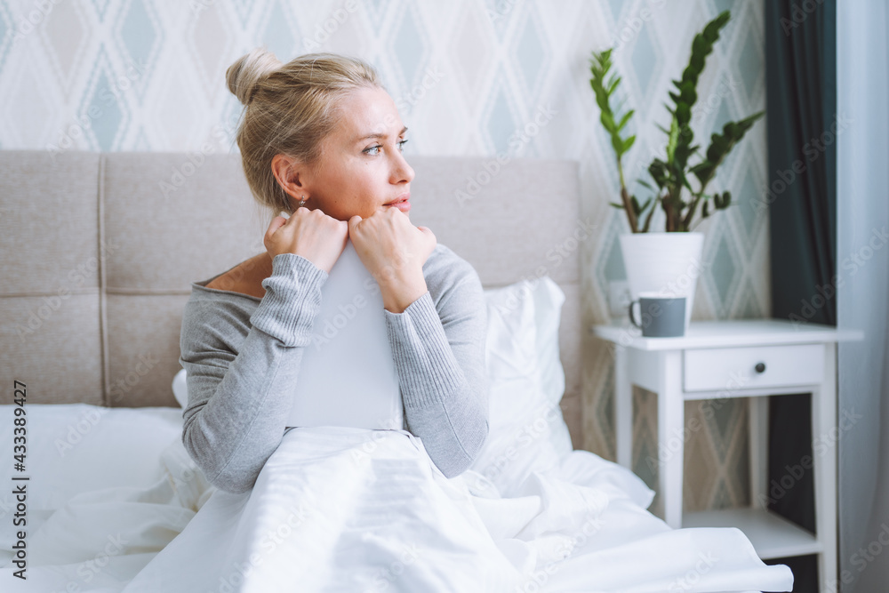 Side view of attractive blonde woman sitting on her bed in light modern bedroom and reading a book with cup of hot beverage Side view of attractive blonde woman sitting on her bed in light modern bedroom and reading a book with cup of hot beverage