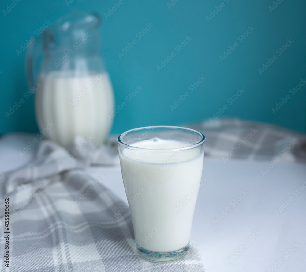 A glass of fresh milk and a jug in the background on a blue background. Healthy and delicious breakfast. Milk Day. copy space for text. selective focus A glass of fresh milk and a jug in the background on a blue background. Healthy and delicious breakfast. Milk Day. copy space for text. selective focus