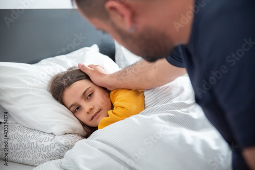 Close-up of sick Caucasian girl lying on bed. Bearded father sitting, stroking daughters head. Single dad taking care of child. Fatherhood, healthcare concept.