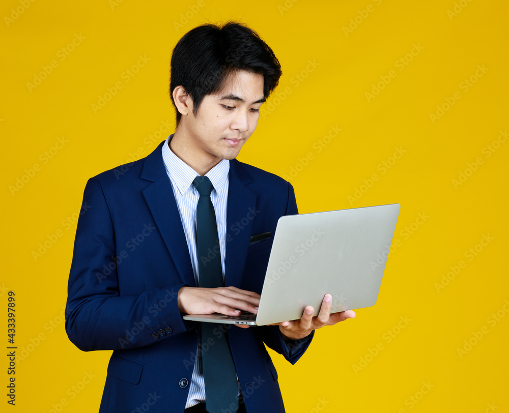 Young Asian businessman in a formal blue suit. Standing work in hand holding laptop. Yellow background gives a modern feel, empty space can enter text.