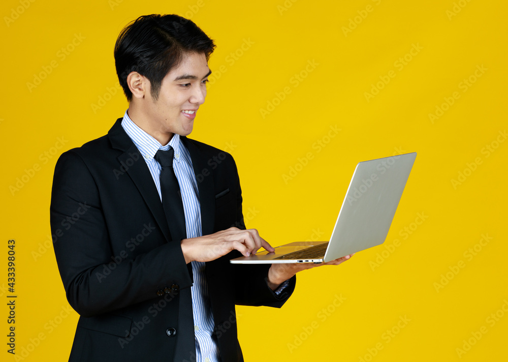 Young Asian businessman in a formal black suit. Standing work in hand holding laptop with smile. Seems he really enjoy his work. Yellow background gives a modern feel, empty space can enter text. Young Asian businessman in a formal black suit. Standing work in hand holding laptop with smile. Seems he really enjoy his work. Yellow background gives a modern feel, empty space can enter text.