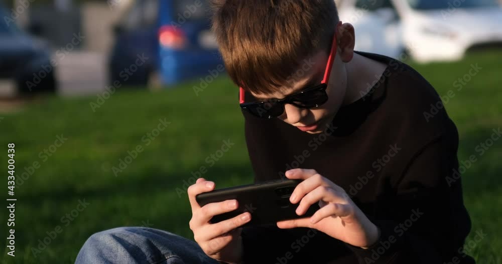 Schoolboy using a smartphone sitting on the lawn. Generation Z. Boy plays on a phone outdoors. Modern technology and children Schoolboy using a smartphone sitting on the lawn. Generation Z. Boy plays on a phone outdoors. Modern technology and children