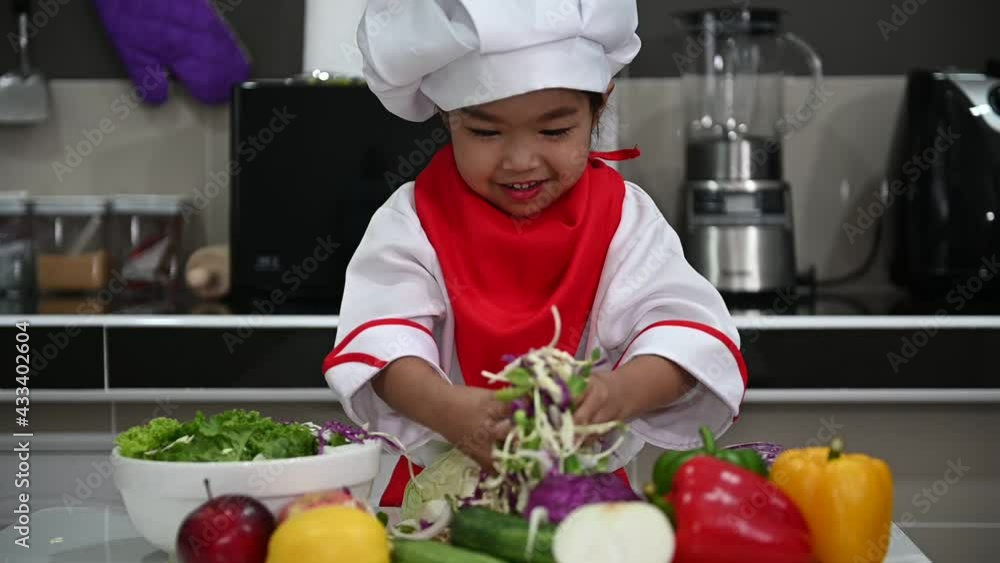 Cute asian girl wear chef uniform with a lot of vegatable on the table in the kitchen room,Make food for eat dinner,Funny time for kids Cute asian girl wear chef uniform with a lot of vegatable on the table in the kitchen room,Make food for eat dinner,Funny time for kids