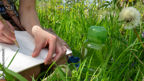 Foto Female hands holding a diary and writing with a pencil in the book, sitting among green grass and flowers in the sunny park