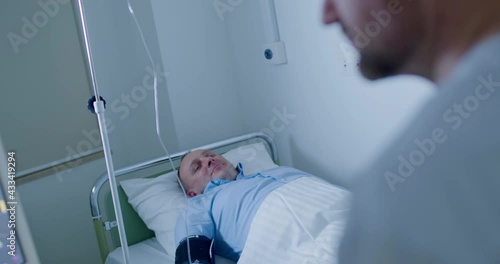 A male patient is laying in the bed at the hospital with a tonometer on his arm and a drip is connecting to his hand while a male doctor is checking a diagram of pulse oxygen saturation on the monitor photo