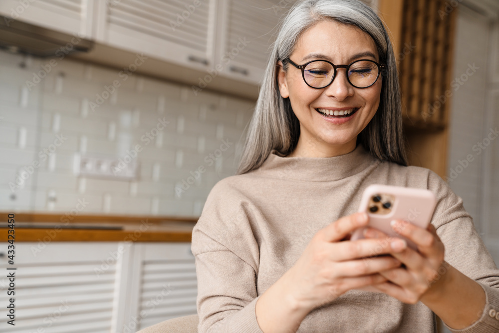 Mature grey woman smiling while using cellphone in kitchen at home