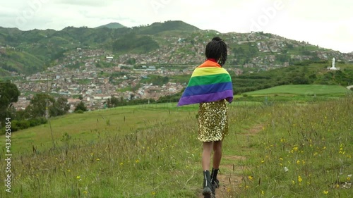Mujer transgénero con vestido dorado y bandera LGTBQ caminando sobre la colina con vista a la ciudad de Cusco