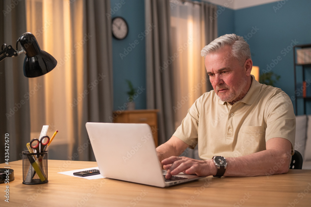 Elderly man sitting at desk with laptop in the evening. Senior works in a salon. Busy man in front of laptop screen. An elderly man shops online, pays bills, browses the news, checks email. Elderly man sitting at desk with laptop in the evening. Senior works in a salon. Busy man in front of laptop screen. An elderly man shops online, pays bills, browses the news, checks email.