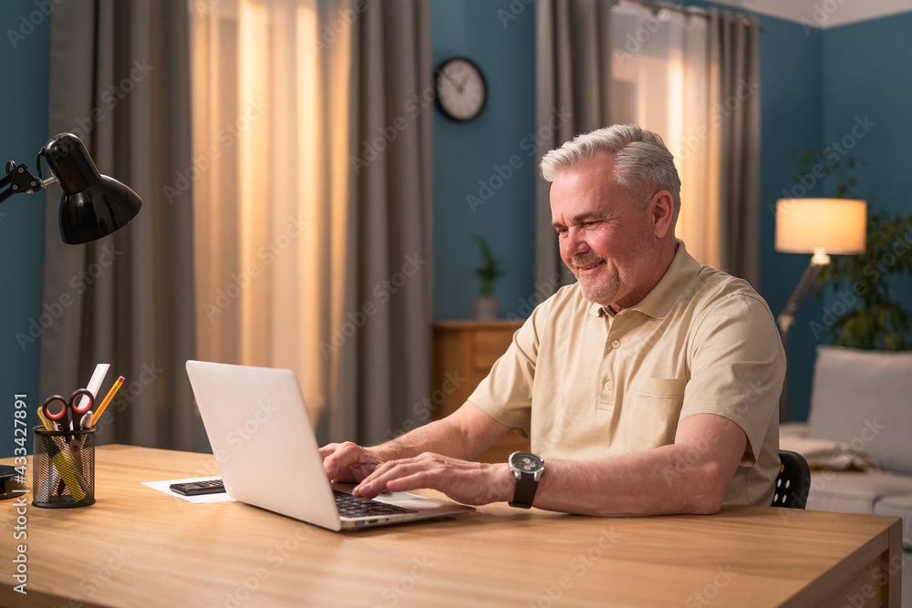 Elderly man sitting at desk with laptop in the evening. Senior works in a salon. Lucky retiree uses a computer at home. Grandfather talks to grandchildren online. A smiling and happy senior talks Elderly man sitting at desk with laptop in the evening. Senior works in a salon. Lucky retiree uses a computer at home. Grandfather talks to grandchildren online. A smiling and happy senior talks