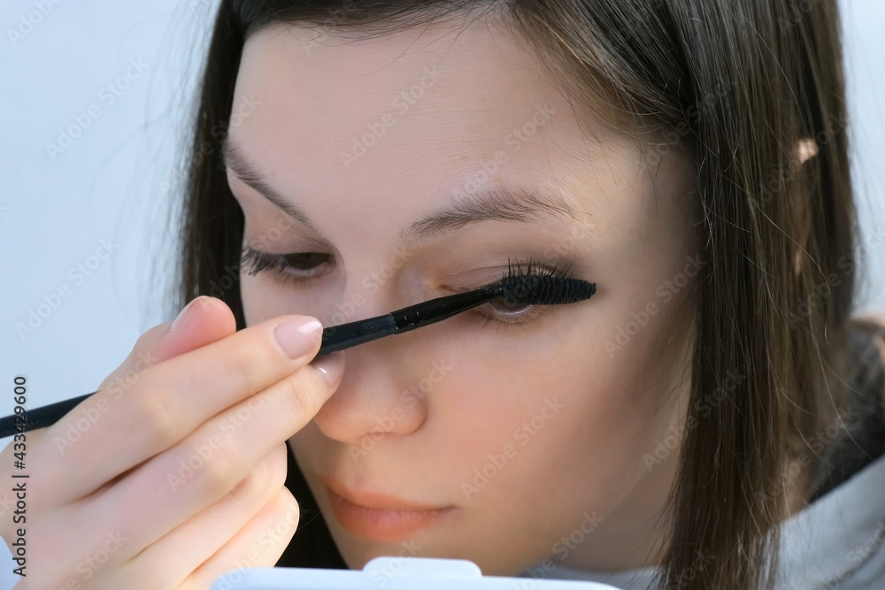 Woman is painting lashes using mascara and brush looking at small mirror, closeup face view. Female is doing makeup. Everyday routine for woman. Woman is painting lashes using mascara and brush looking at small mirror, closeup face view. Female is doing makeup. Everyday routine for woman.