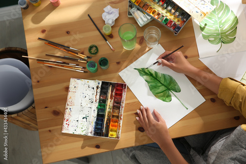 Billede på lærred Young woman drawing leaf with watercolors at table indoors, top view