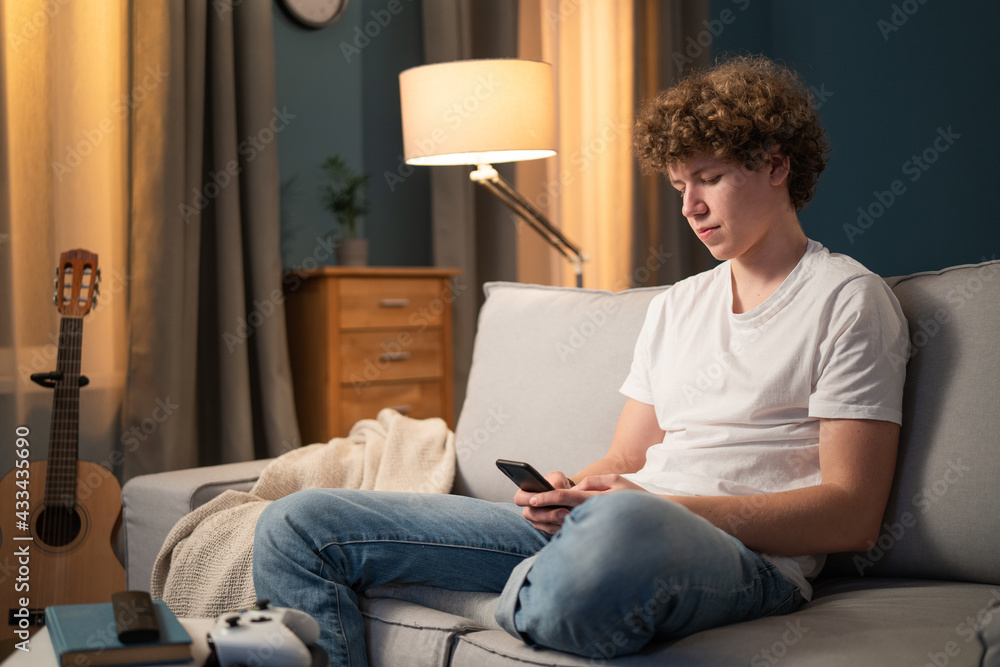 A bored young boy, teenager, schoolboy, student is relaxing on the couch in the evening with his phone, smartphone in hand. The young boy sends messages to friends, browses social media