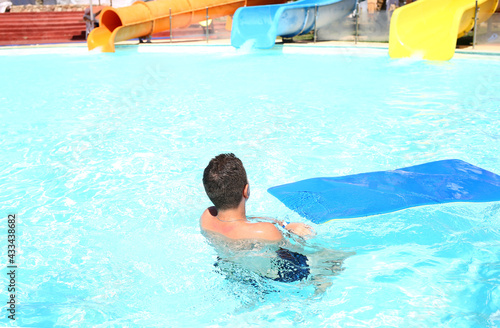 A teenage boy back view in the pool with a soft mat for descending from the water attraction. Entertainment for children. Recreation and tourism concept. Blue water. Copy space