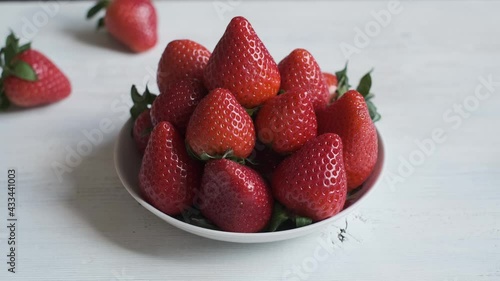 White plate of red ripe strawberries on a wooden white wooden background. In the background, a strawberry berry rolls past on the table.