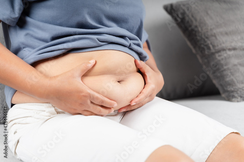 Close up of chubby woman holding her belly fat on the sofa. Diet lifestyle to reduce belly and shape up healthy abdominal muscle concept.