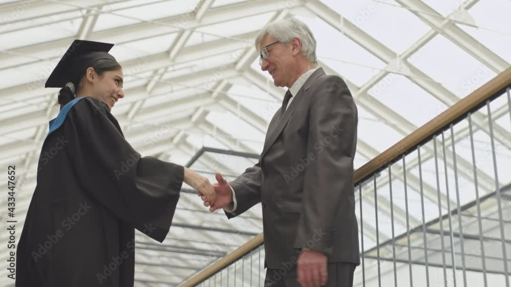 Low angle view of aged male university tutor shaking hand of young mixed-race female student wearing university graduate gown and hat. Then they making selfie together using smartphone Low angle view of aged male university tutor shaking hand of young mixed-race female student wearing university graduate gown and hat. Then they making selfie together using smartphone
