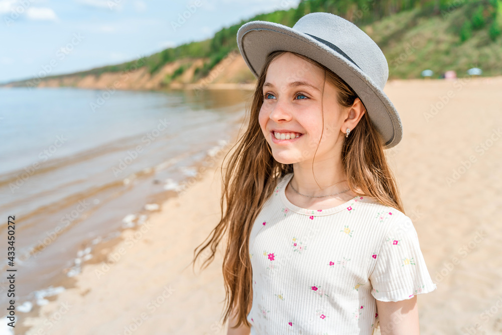Portrait of a smiling little girl in a wide-brimmed hat standing on a sandy beach by the sea