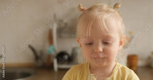 A little blonde girl drinks water from a glass in the kitchen looking at the camera and smiling