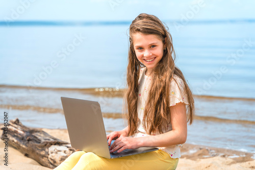 A little girl with long hair sits on a log on the beach with a laptop on her lap. The child uses a laptop