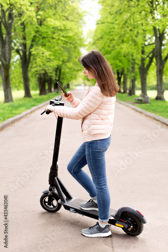 Young beautiful girl in  riding in the Park on an electric scooter on a summer day  uses phone. Walk in the Park 