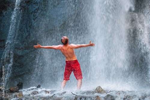Shirtless Man dressed only red trekking shorts standing under the mountain river waterfall, spread his arms and enjoying the splashing Nature power. Traveling, trekking and nature concept image.