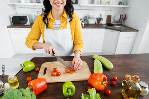 cropped view of cheerful woman cutting cherry tomatoes on wooden chopping board near vegetables