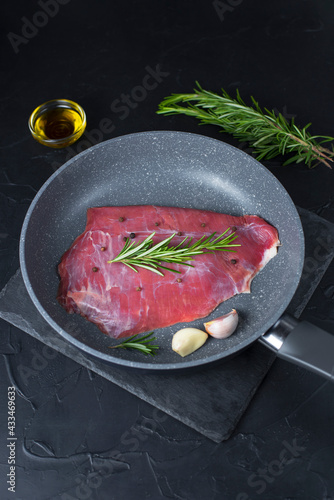 Fresh raw flank steak beef in the pan with rosemary, garlic, olive oil on dark background. Classic marinades recipe for roasting or grilling. Meat food menu