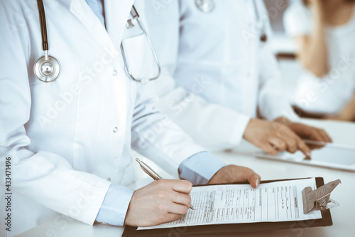 Unknown woman-doctors at work with patient at the background. Female physicians filling up medical documents or prescription while standing in hospital reception desk, close-up. Health care concept