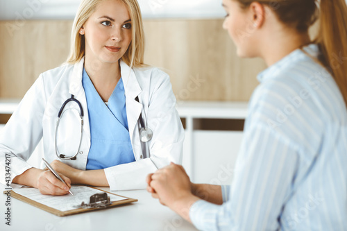 Woman-doctor and patient sitting and talking in hospital or clinic. Blonde therapist is cheerfully smiling. Medicine concept