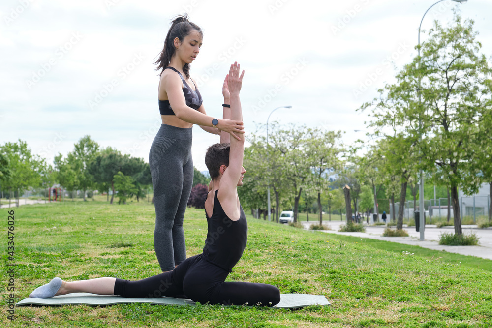 Fototapeta premium Female personal trainer helping young man in a park with arms up pigeon yoga pose. Stretching exercises on a mat outside.